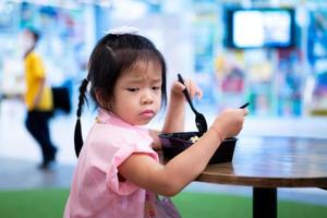 asiatico carino ragazza è nel cattivo umore mentre mangiare pranzo o cena. bambino accigliato. 5 anni vecchio scuola materna ragazzo è Affamato. foto