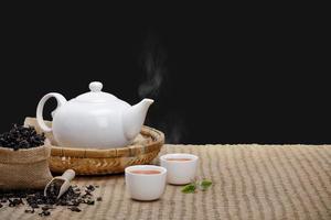 caldo tazza di tè con teiera, verde tè le foglie e secco erbe aromatiche su il bambù stuoia a mattina isolato nero sfondo con vuoto spazio, biologico Prodotto a partire dal il natura per salutare con tradizionale foto