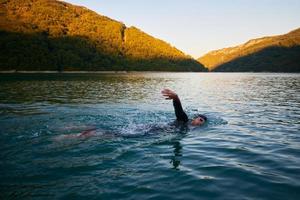 atleta di triathlon che nuota sul lago all'alba indossando la muta foto