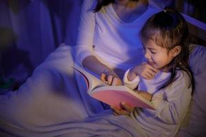 mamma lettura un' libro con sua figlia su letto nel un' buio Camera da letto a notte prima dormire, paternità e contento momenti concetto foto