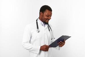 sorridente nero barbuto medico uomo nel bianca accappatoio con stetoscopio sembra per medico grafico su appunti foto