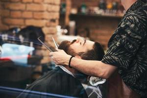 barbuto maschio seduta nel un poltrona nel un' barbiere negozio mentre parrucchiere viaggio il suo barba con forbici. avvicinamento di barbiere tosatura barba per uomo nel barbiere foto