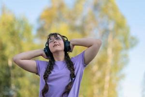 un' adolescente ragazza sta su il strada, presse cuffie per sua orecchie con sua mani e ascolta per musica con sua occhi Chiuso. foto