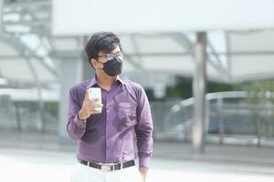 sudorazione asiatico giovane uomo d'affari con chirurgico viso maschera utilizzando mobile Telefono durante ufficio ore come parte di attività commerciale nuovo normale foto