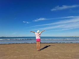 ragazza con cappello apertura sua braccia nel davanti di il mare, su un' sabbioso spiaggia. foto