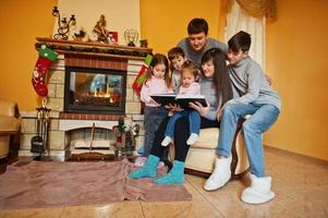 felice giovane famiglia numerosa accanto a un camino nel caldo soggiorno in una giornata invernale. madre con quattro bambini a casa legge il libro. foto