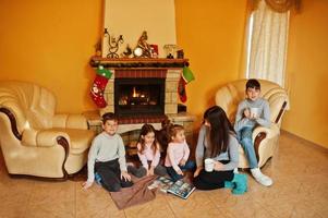 felice giovane famiglia numerosa accanto a un camino nel caldo soggiorno in una giornata invernale. madre con quattro bambini a casa legge il libro. foto