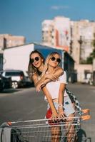 Due giovane ragazze nel occhiali da sole in posa per il telecamera su il auto parcheggio. foto