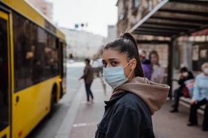 giovane donna indossare chirurgico maschera all'aperto a autobus fermare nel il strada foto