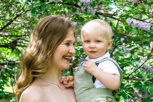 ritratto di giovane sorridente madre con sua poco figlio nel luce del sole su sfondo di fioritura lilla alberi. foto