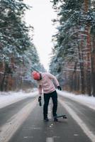 skateboarder in piedi su il strada nel il mezzo di il foresta, circondato di neve foto