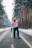 skateboarder in piedi su il strada nel il mezzo di il foresta, circondato di neve foto