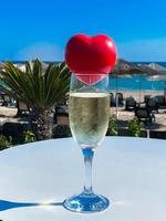 un' rosso cuore bugie su il bicchiere di Champagne con il mare e palme a il sfondo foto