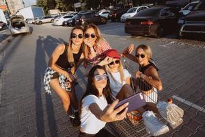 cinque giovane ragazze avere divertimento con un' supermercato carrello su un' auto parco foto