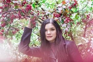 ritratto di attraente donna con lungo Marrone capelli nel pelle giacca nel giardino con fiorire rosso Mela albero fiori e gocce di pioggia. foto