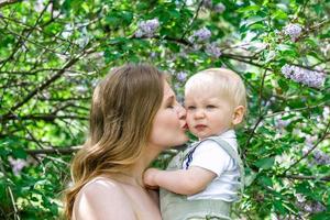 giovane bellissimo madre è baci sua poco figlio nel luce del sole su sfondo di fioritura lilla alberi. foto