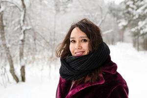 bellissimo sorridente donna con lungo Marrone capelli nel un' cappotto a partire dal finto pelliccia e grande nero sciarpa su un' sfondo di inverno foresta. foto