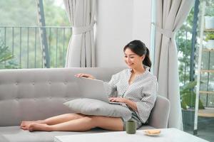 un' asiatico donna la spesa tempo a casa, seduta su divano, utilizzando sua il computer portatile foto