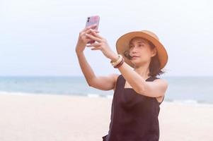 un asiatico donna sta per prendere un' autoscatto con sua mobile Telefono nel il sabbia durante un' estate vacanza su il spiaggia foto