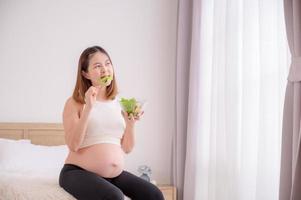 un asiatico donna si siede nel sua Camera da letto mangiare sano, biologico insalate mentre lei è incinta fino a il tempo di sua del bambino nascita foto