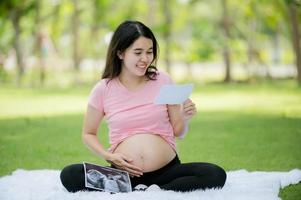 un asiatico prima volta incinta donna si siede nel il giardino guardare a il immagini a partire dal il ospedale ultrasuono per dai un'occhiata il Salute di sua in crescita bambino foto