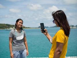 un' donna nel un' giallo camicia è Tenere un' Telefono per prendere immagini di un' ragazza nel un' grigio camicia. con un' sfondo di blu acqua e piccolo montagne. esso è turismo dopo il corona virus scoppio. foto