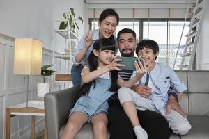 contento asiatico tailandese genitori, e bambini in linea video chiamata con famiglia attraverso Internet su divano nel casa vivente camera, bello fine settimana, e domestico benessere stile di vita con Internet tecnologia mobile Telefono. foto