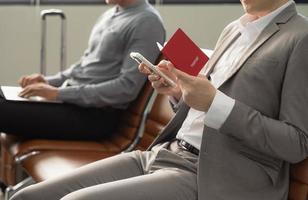 uomo d'affari Tenere passaporto seduta a aeroporto terminale e controllo per il suo volo su mobile Telefono, in attesa per aereo e Lavorando mentre in viaggio foto