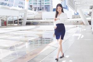 giovane attività commerciale asiatico donna è a piedi per partire per il ufficio o posto di lavoro quale lei Guarda a polso orologio nel il grande città con attività commerciale edifici come un' sfondo. foto