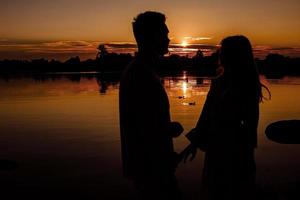 silhouette di amorevole coppia abbracciare su il lago a tramonto. bellissimo giovane coppia nel amore a piedi su il riva di il lago a tramonto nel il raggi di luminosa luce. copia spazio. selettivo messa a fuoco. foto