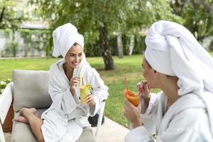 Due amichevole donne nel bianca accappatoi seduta nel terme con bevande e parlando per ogni altro dopo terme cura routine foto