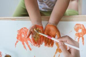 madre pittura su mano sua asiatico bambino ragazza sorridente pittura con colorato vernici utilizzando acquerello. asiatico ragazza utilizzando pennello disegno colore. bambino attività stile di vita concetto. foto