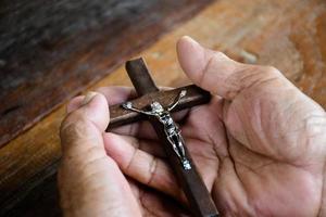 avvicinamento di legno attraversare quale ha un' metallo statua di crocifisso Gesù è nel il mani di un asiatico anziano cattolico mentre preghiere nel un' Locale Chiesa, morbido e selettivo messa a fuoco. foto