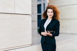 giovane imprenditrice di successo in abiti eleganti, con capelli lussureggianti, tenendo il tablet in mano mentre va al lavoro. splendida femmina che tiene computer tablet digitale. tecnologia e affari foto