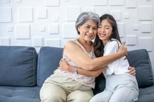 asiatico nonna e nipotina abbracciato con contento umore su il divano nel casa. foto