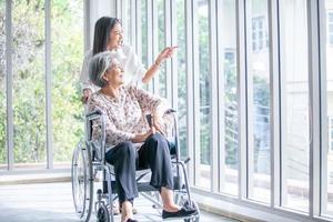 bellissimo asiatico ragazza Aiuto e prendere cura di anziano anziano donna seduta su sedia a rotelle a vivente camera nel Casa, anziano terapia paziente a casa concetto. foto