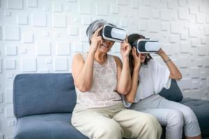 asiatico nonna e nipotina utilizzando virtuale la realtà giocare Giochi a casa. anziano e giovane donna indossare vr-headset bicchieri su divano nel vivente camera. foto