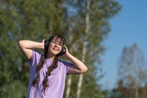 un' adolescente ragazza sta su il strada, presse cuffie per sua orecchie con sua mani e ascolta per musica con sua occhi Chiuso. foto
