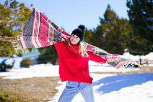 felice giovane donna bionda che ondeggia la sua sciarpa nel vento in una foresta tra le montagne innevate. foto