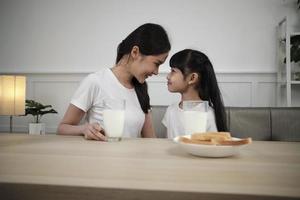 salutare asiatico tailandese famiglia, contento poco figlia, e giovane madre viso per viso a cenare tavolo con fresco latte nel bicchiere e pane cura insieme, benessere nutrizione prima colazione pasto, mattina stile di vita. foto