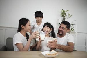 un' salutare asiatico tailandese famiglia, poco figli, e giovane genitori bevanda fresco bianca latte nel bicchiere e pane gioia insieme a un' cenare tavolo nel mattina, benessere nutrizione casa prima colazione pasto stile di vita. foto