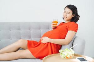 bella giovane asiatico incinta donna mangiare insalata e potabile latte quando seduta su divano. foto