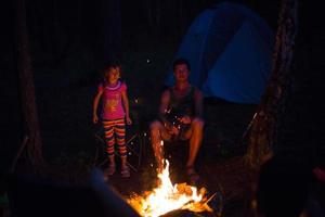 papà e figlia si siedono di notte accanto al fuoco all'aria aperta in estate nella natura. campeggio in famiglia, incontri intorno al fuoco. festa del papà, barbecue. lanterna e tenda da campeggio foto