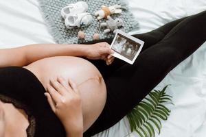 mani della donna incinta che tengono sulla pancia facendo un simbolo di cura e amore sulla camera da letto con l'immagine ad ultrasuoni. gestante in attesa e preparazione per la nascita del bambino. concetto di donna incinta. foto