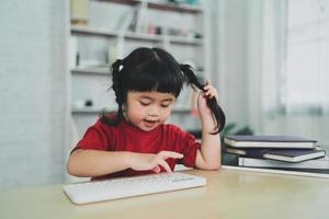 asiatico bambino ragazza indossare un' rosso maglietta uso senza fili tastiera e studia in linea su legna tavolo scrivania nel soggiorno a casa. formazione scolastica apprendimento in linea a partire dal casa concetto. foto