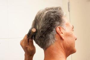uomo maturo oltre i 60 anni che si pettina i capelli per lo più grigi nella parte posteriore della testa davanti a uno specchio foto