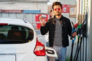 uomo del sud asiatico o maschio indiano che fa rifornimento di carburante alla sua macchina bianca sulla stazione di servizio e tiene la carta di credito. foto