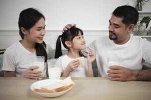 un' salutare asiatico tailandese famiglia, un' poco figlia, e giovane genitori bevanda fresco bianca latte nel bicchiere e pane gioia insieme a un' cenare tavolo nel mattina, benessere nutrizione casa prima colazione pasto stile di vita. foto