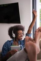 africano americano donna a casa nel sedia con tavoletta e testa telefoni foto