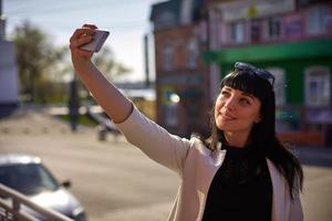 carino donna con occhiali da sole e lungo capelli è utilizzando mobile dispositivo godendo raggi di sole e caldo giorno all'aperto. ritratto di sorridente brunetta fare autoscatto su un' smartphone foto